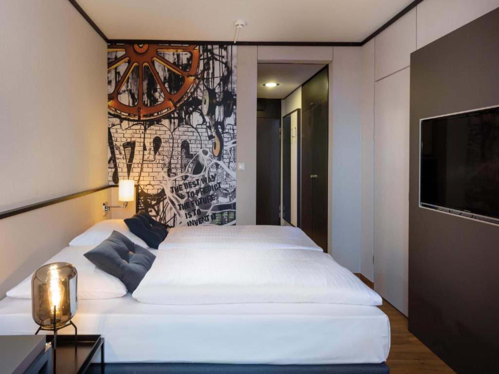 A modern hotel room with a large bed, a wall mural, and a TV.