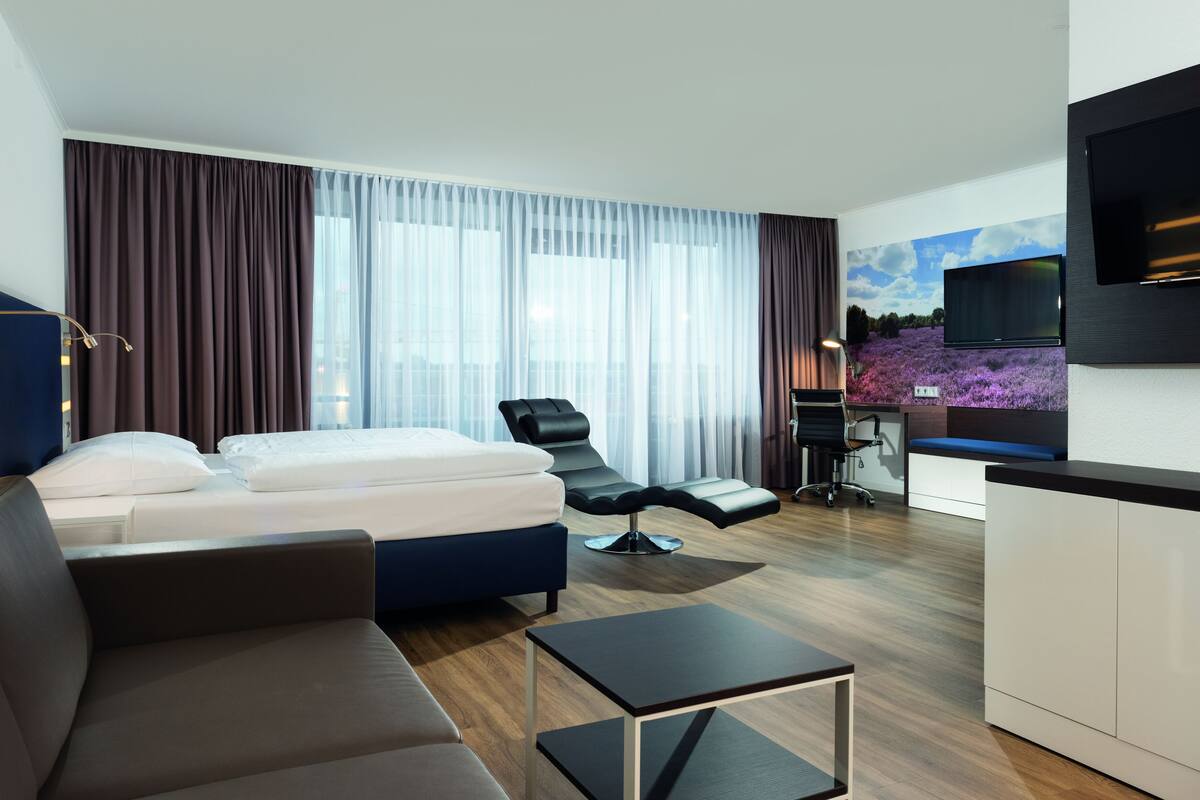 A modern hotel room with a bed, a sofa, a desk, a chair, and a TV.