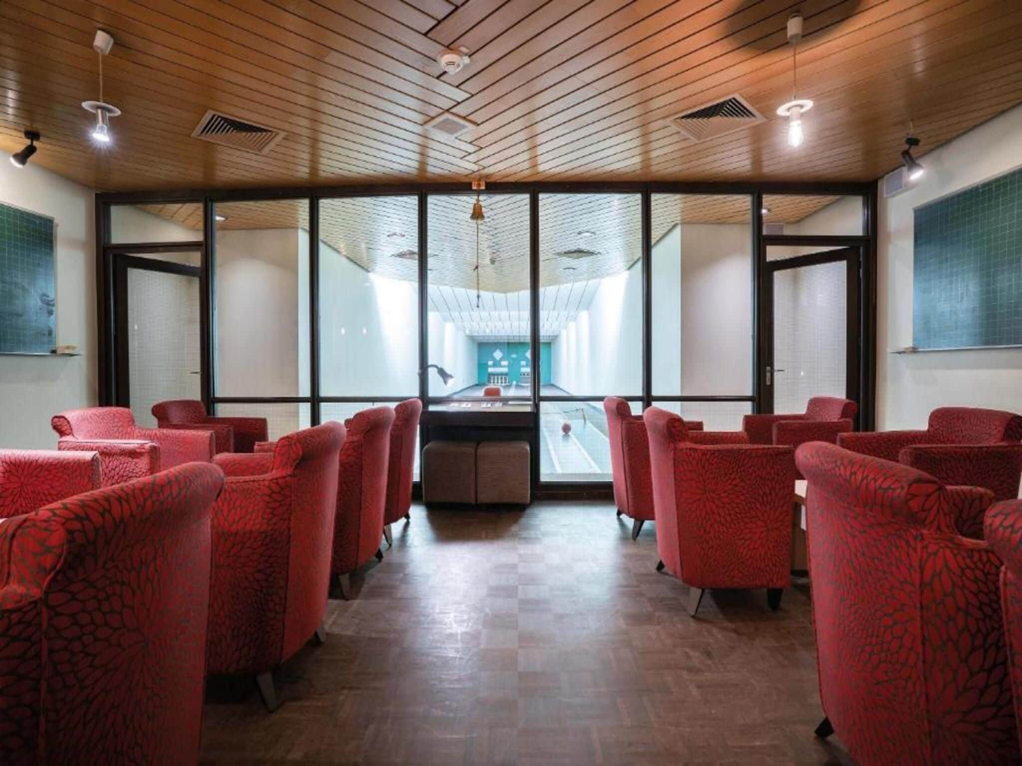 A room with red upholstered chairs arranged in rows, large windows, and a wooden ceiling.