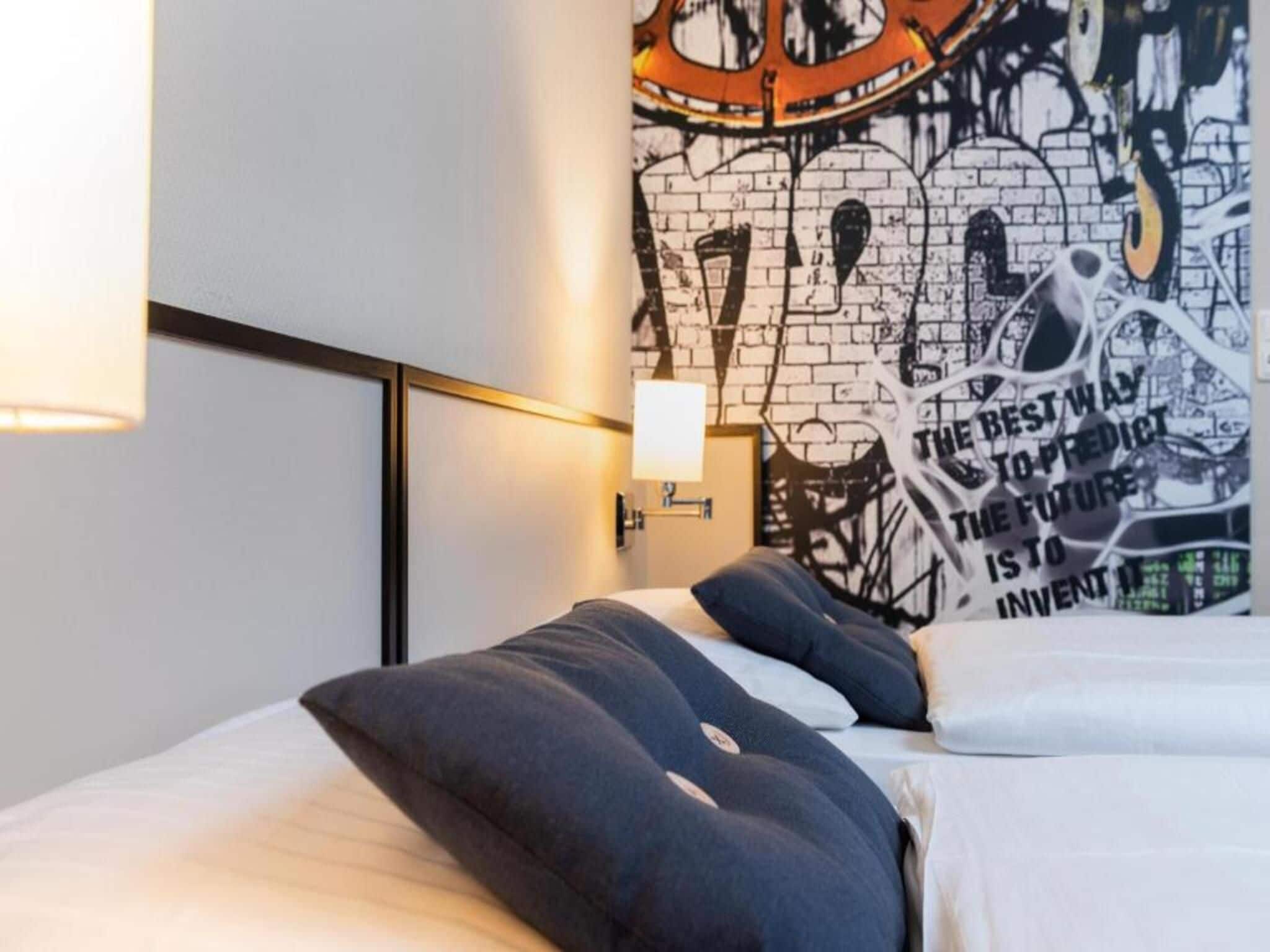 A modern hotel room with a bed, pillows, a wall with graffiti art, and a nightstand with a lamp.