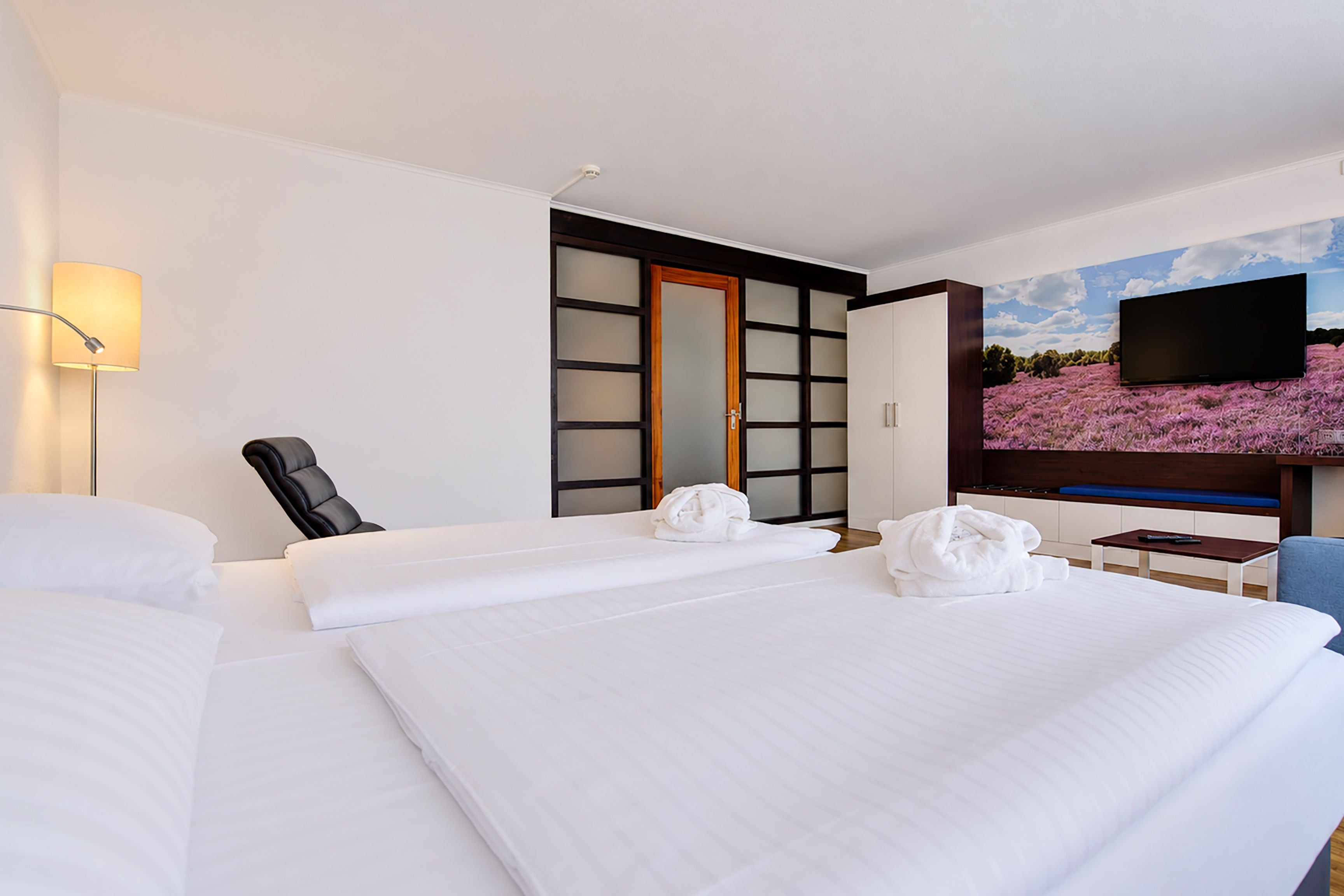 A hotel room with a bed, a chair, a desk, a flat-screen TV, and a painting of a landscape.