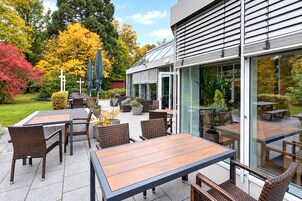 An outdoor patio area with tables and chairs, a glass door entrance, and a garden with trees and shrubs.