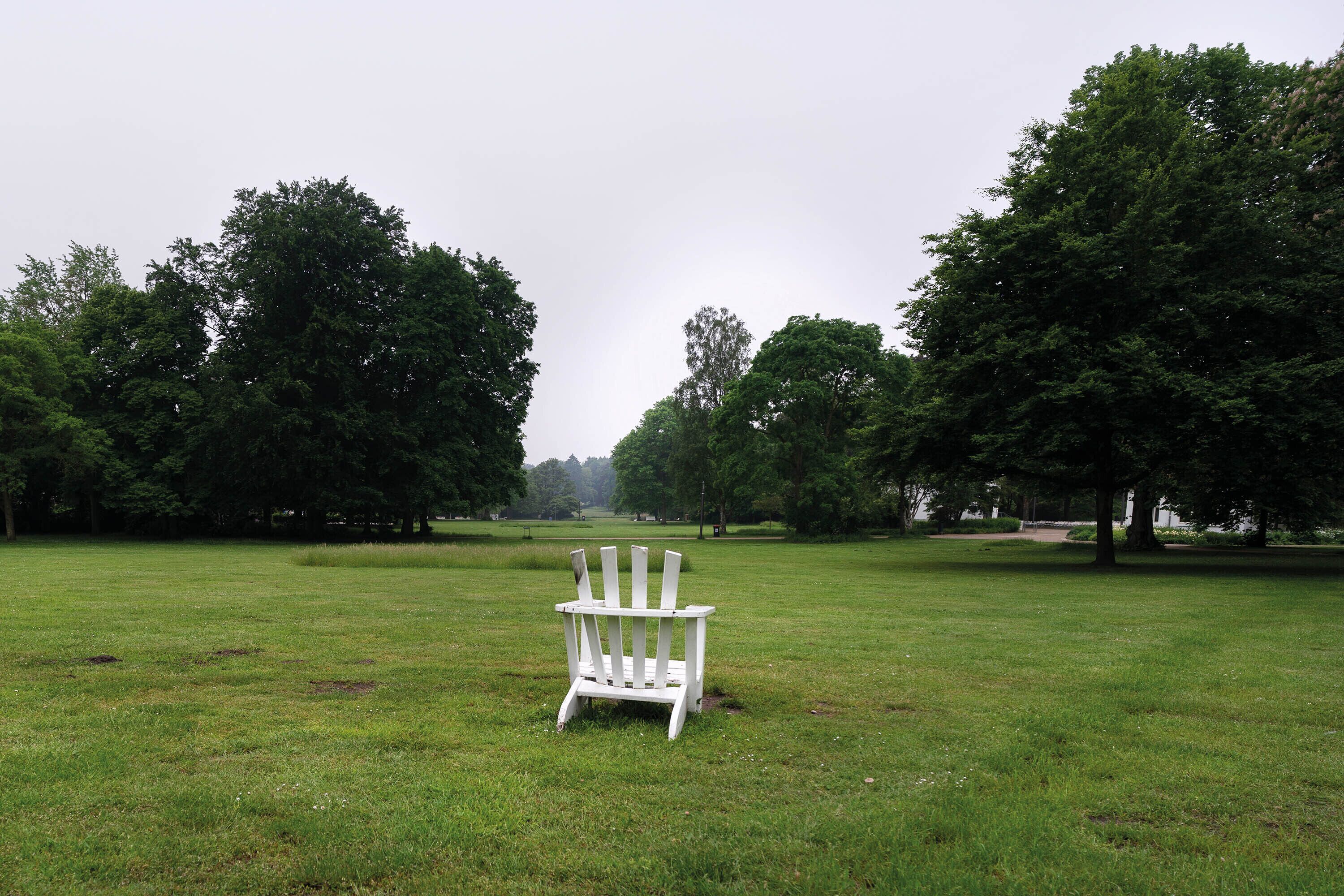 A white Adirondack chair in a grassy field with trees in the background.