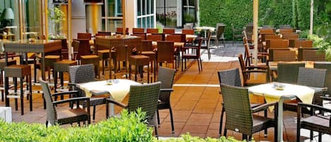 An outdoor dining area with multiple tables and chairs, yellow umbrellas, and a garden with various plants and flowers.