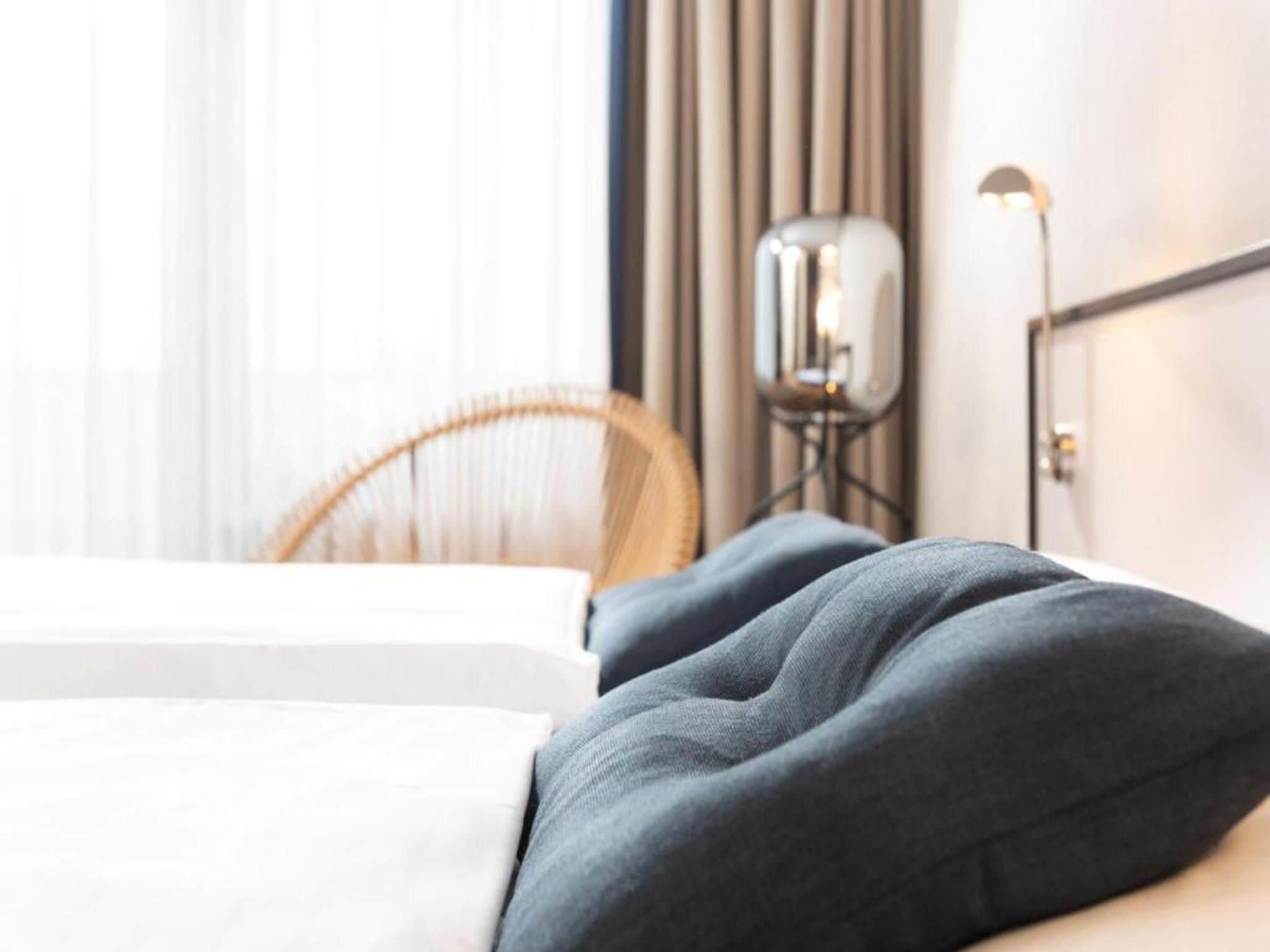 A bed with a white sheet and a dark gray pillow, a wicker chair, and a floor lamp in a hotel room.