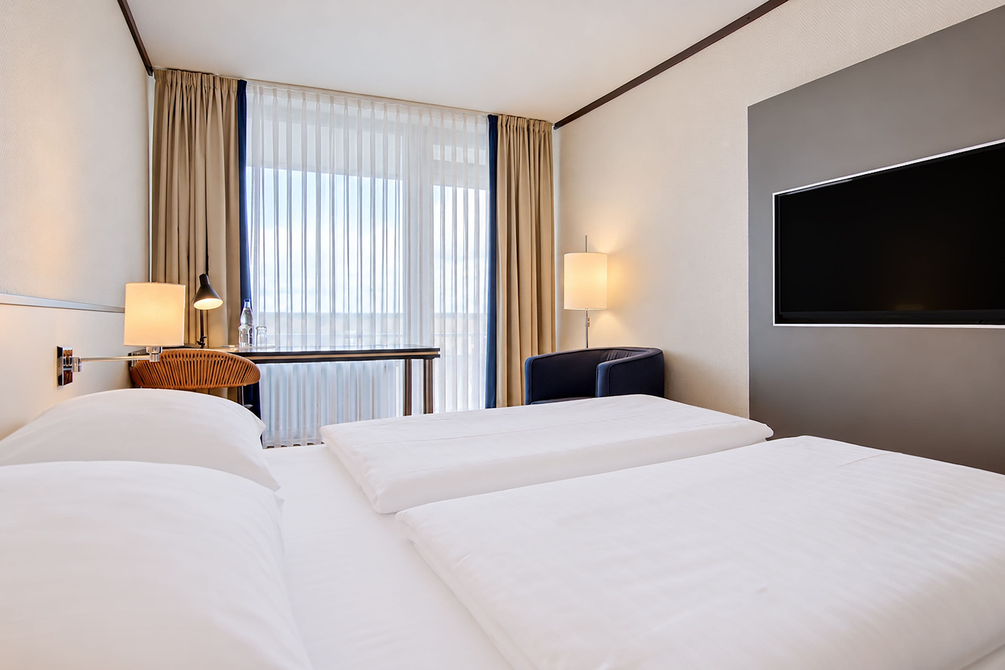 Una camera d'albergo con due letti, una scrivania, una sedia, una televisione e una finestra con tende.