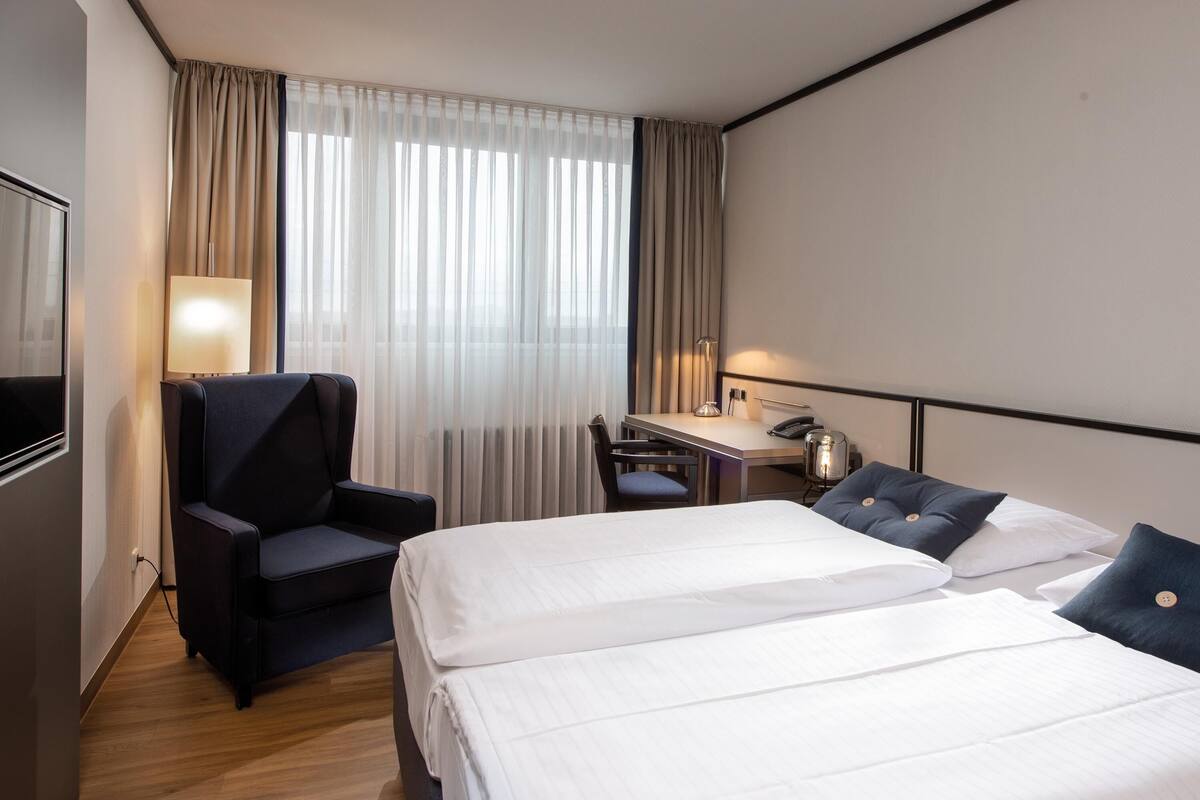 A hotel room with two beds, a desk, a chair, a lamp, and a TV.