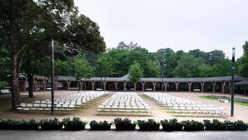 An outdoor seating area with rows of white chairs and tables, surrounded by trees and covered structures.