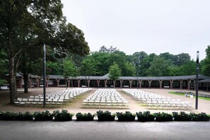 An outdoor seating area with rows of white chairs and tables, surrounded by trees and covered structures.