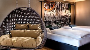 Une chambre moderne avec un fauteuil suspendu, un lit recouvert de linge de maison blanc et une œuvre d’art murale.