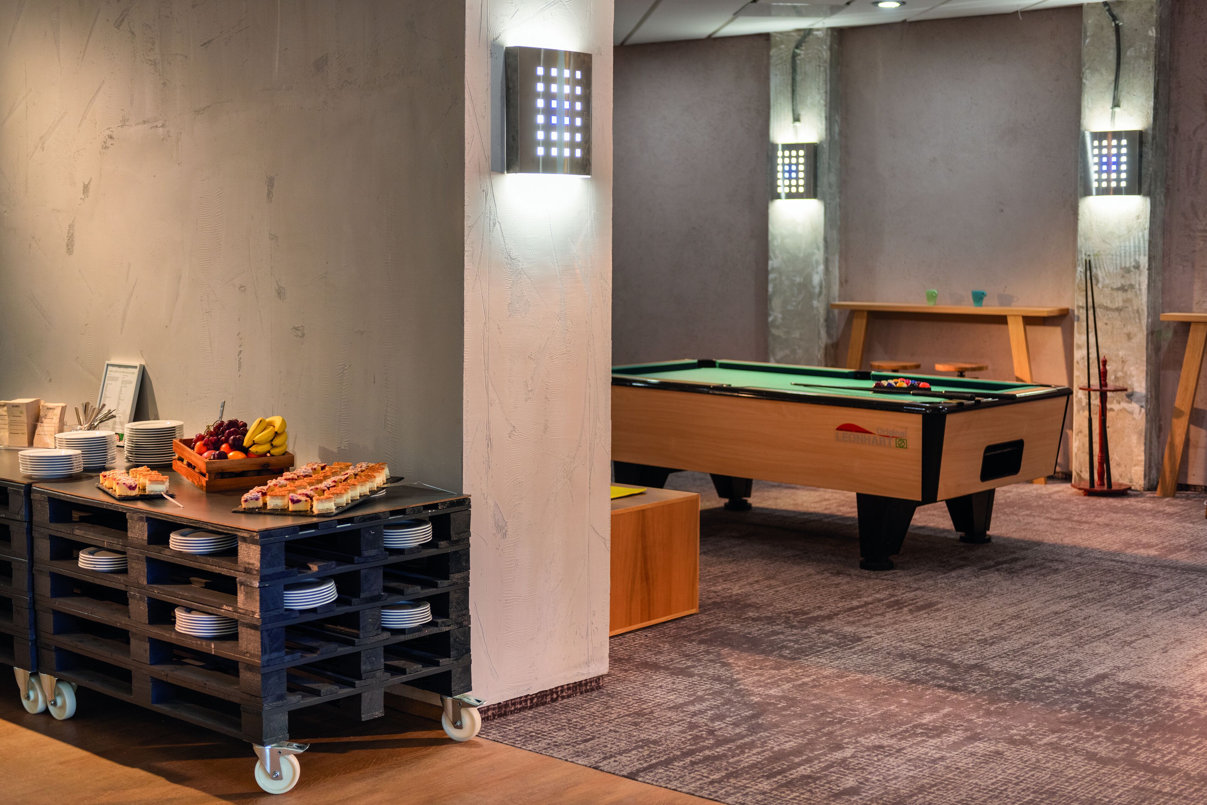Una mesa de billar en una habitación con piso de madera, un carrito de snacks con platos y fruta, e iluminación en las paredes.