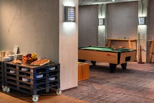 A pool table in a room with a wooden floor, a snack cart with plates and fruit, and wall-mounted lights.