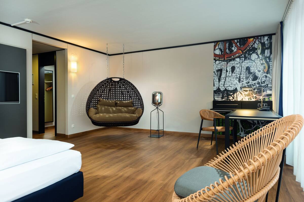 A modern hotel room with a wooden floor, a hanging chair, a dining table, and a large wall mural.