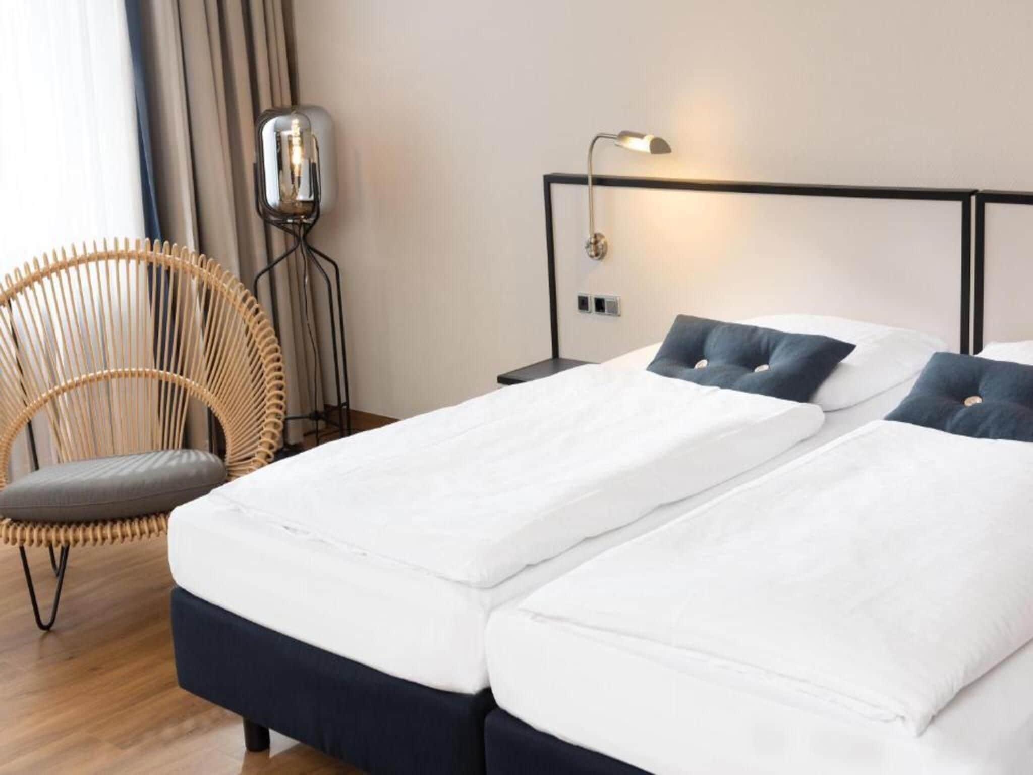 A double bed with white linens and dark blue headboard, a wicker chair with a grey cushion, and a bedside table with a lamp.