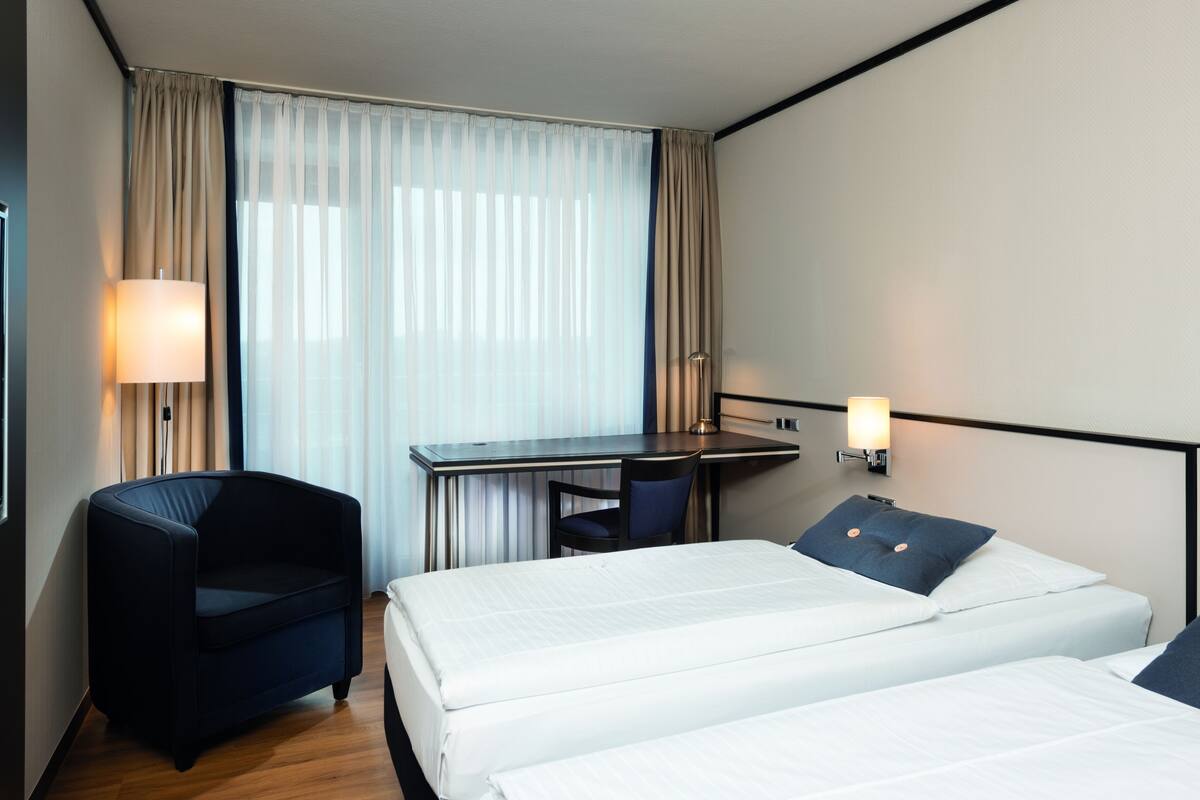 A hotel room with two beds, a desk, a chair, and two wall-mounted lamps.