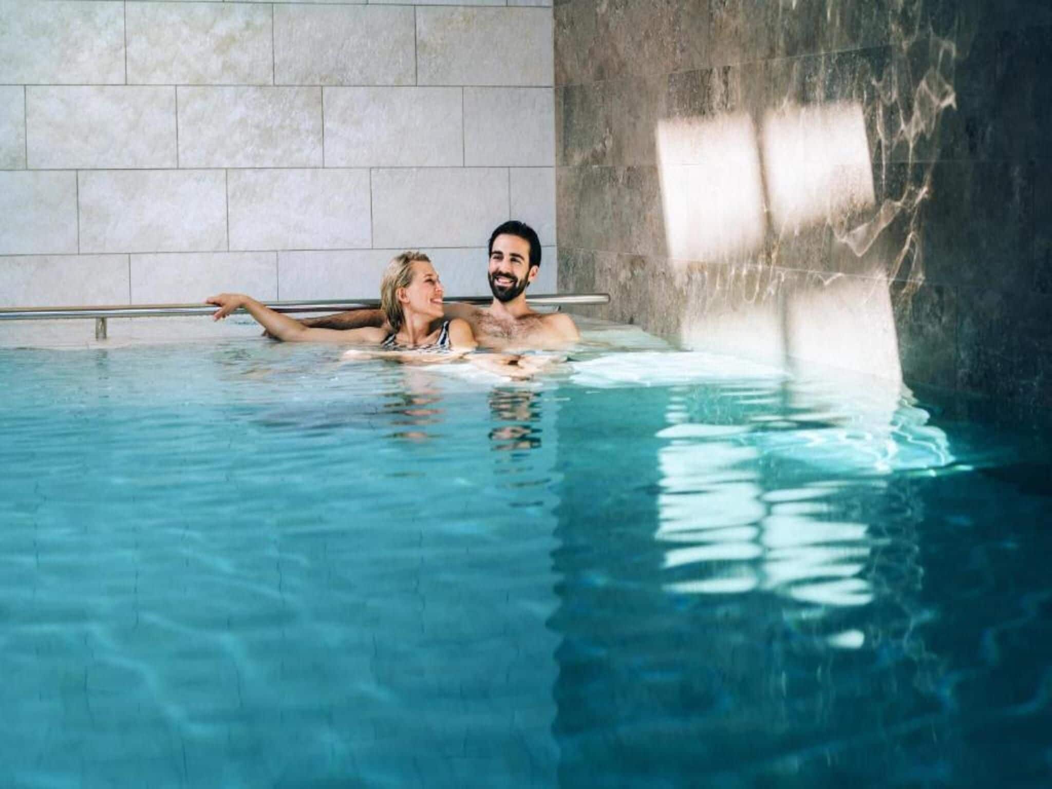 Una pareja en una piscina interior transparente con pared de azulejos y barandilla de metal.