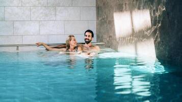 A couple in a clear indoor swimming pool with a tiled wall and a metal railing.