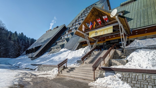 A modern building with a sloped roof and multiple windows. There are stairs leading up to the entrance, and a sign with flags is visible. - Pinia Hotel & Resort (Spindleruv Mlyn)