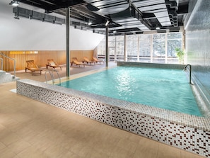 An indoor swimming pool with tiled walls, wooden loungers, and a view of trees outside.