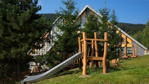 A playground with a slide and climbing structures, surrounded by trees and a building with a sloped roof. - Pinia Hotel & Resort (Spindleruv Mlyn)