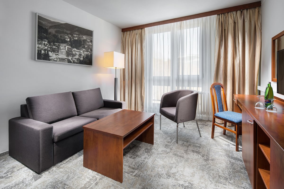 Ein modernes Hotelzimmer mit grauem Sofa, einem Holztisch, einem blauen Stuhl mit gemustertem Kissen, einem Schreibtisch aus Holz mit Flasche und Gläsern sowie einem großen Fenster mit transparenten Vorhängen.
