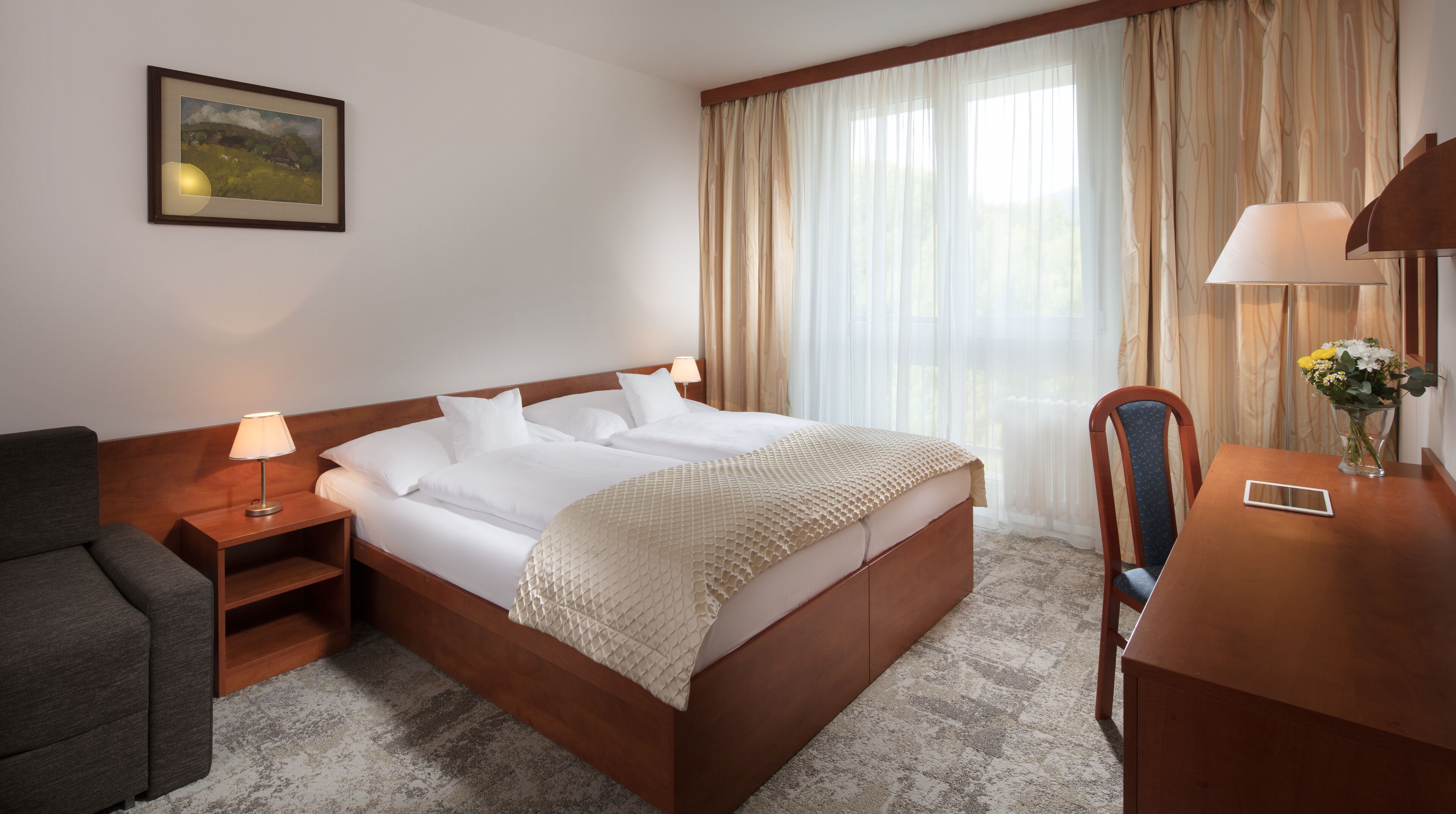 Habitación de hotel con cama doble, mesita de noche con lámpara, una silla, escritorio con un jarrón de flores y un cuadro en la pared.