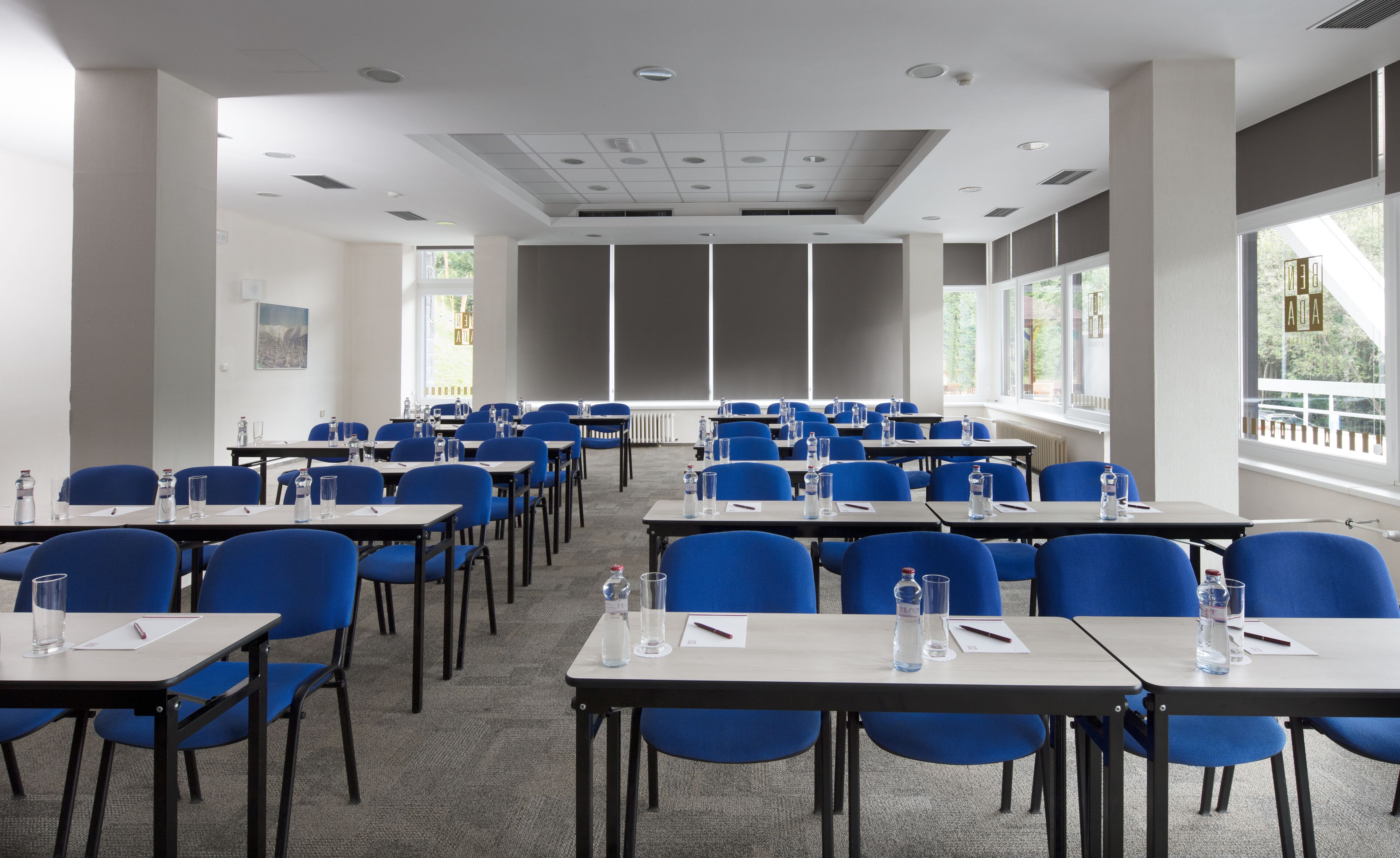 A conference room with rows of blue chairs and tables, equipped with water bottles and pens, large windows, and a white ceiling with recessed lighting.