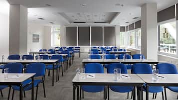 Un salón de conferencias con filas de sillas y mesas azules, equipado con botellas de agua y bolígrafos, ventanas amplias y un techo blanco con iluminación empotrada.