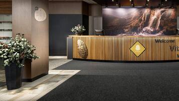 A reception desk with a wooden finish, a large screen displaying a waterfall, and potted plants on either side.