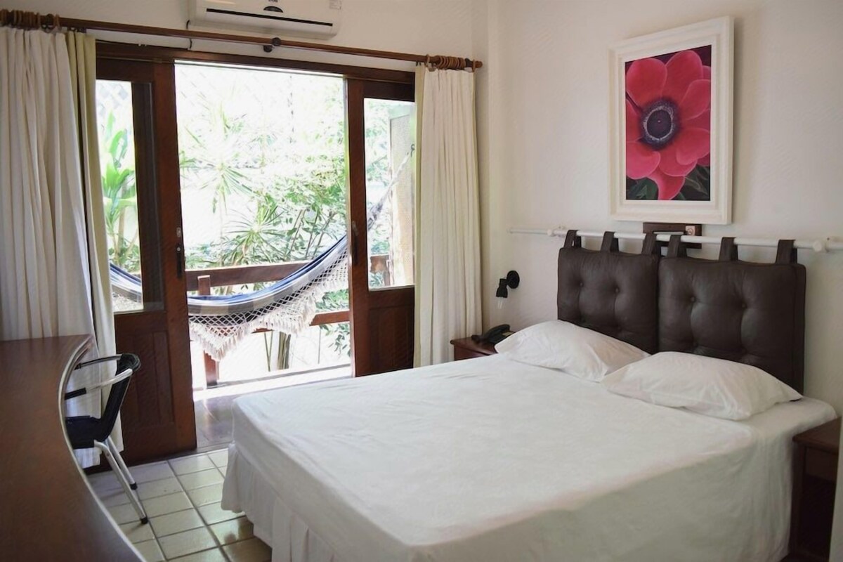 Une chambre d’hôtel avec un lit, un bureau, une chaise, un hamac et une grande fenêtre avec des rideaux.