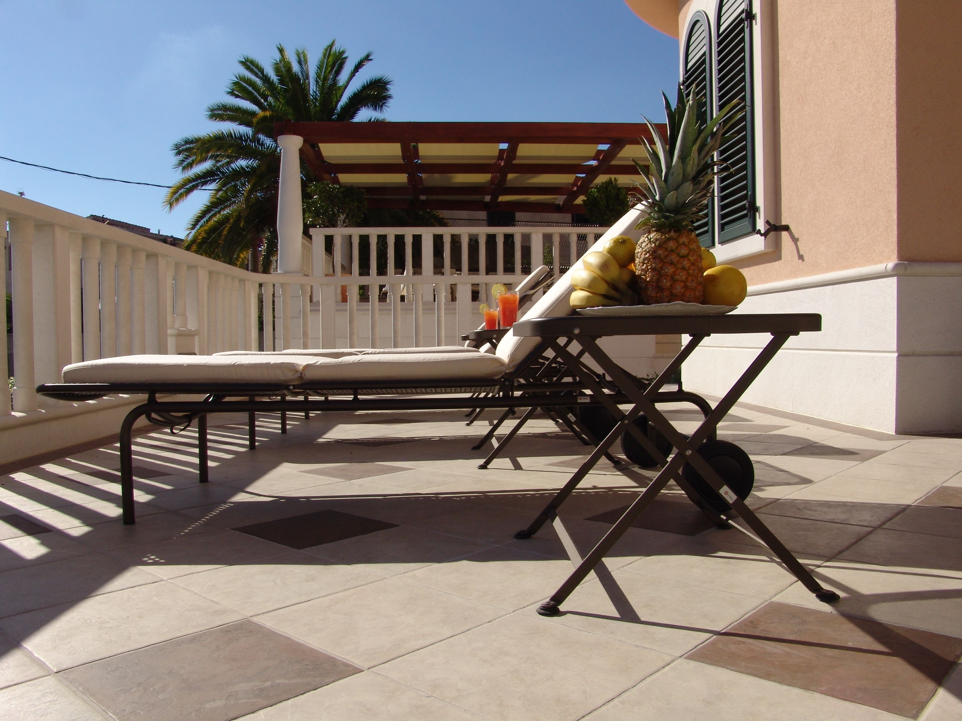 A sun lounger with a table and a pineapple on it, a white fence, and a building with a red roof in the background.