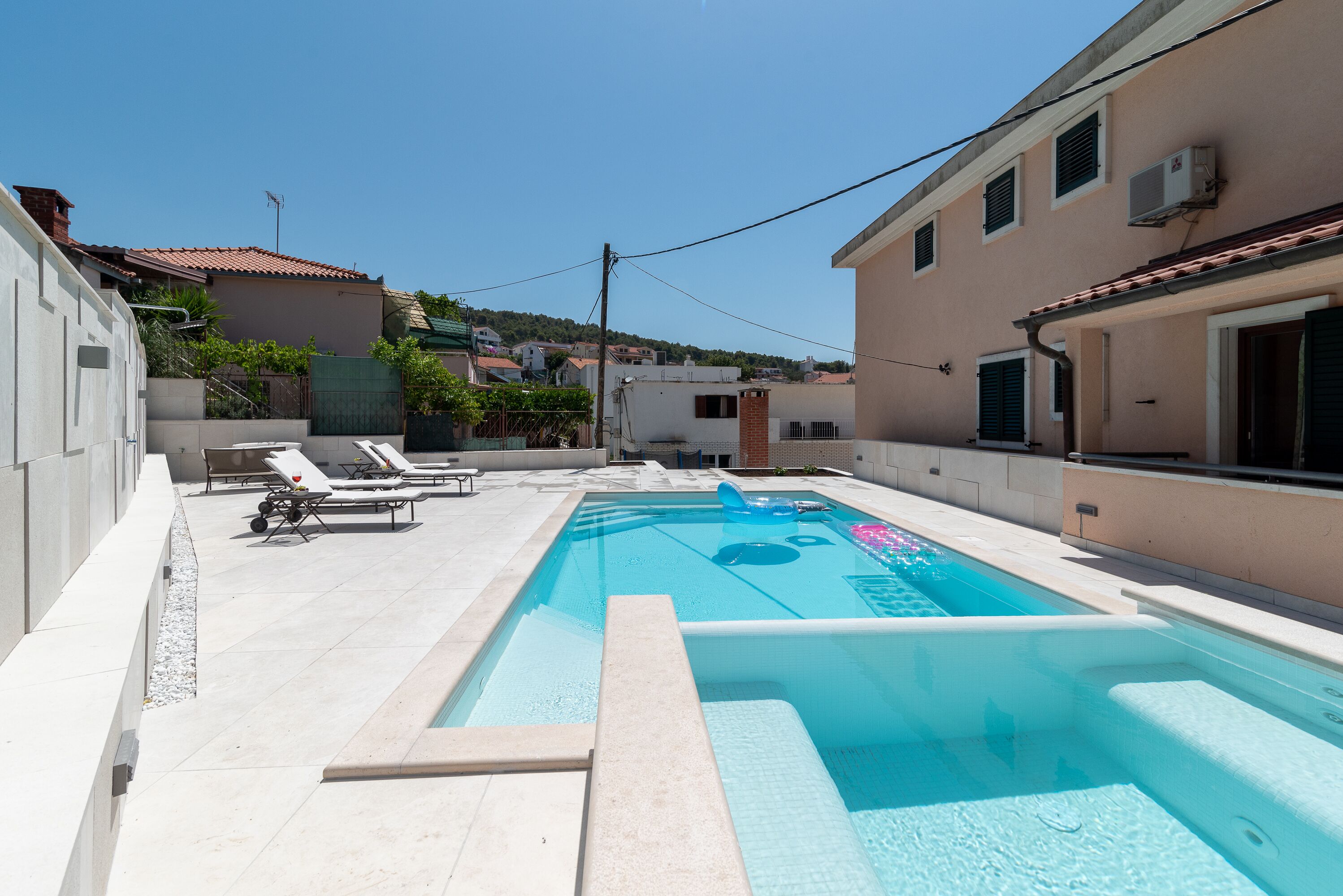 Una piscina con sillas de descanso y una tumbona, rodeada de edificios y un cielo despejado.