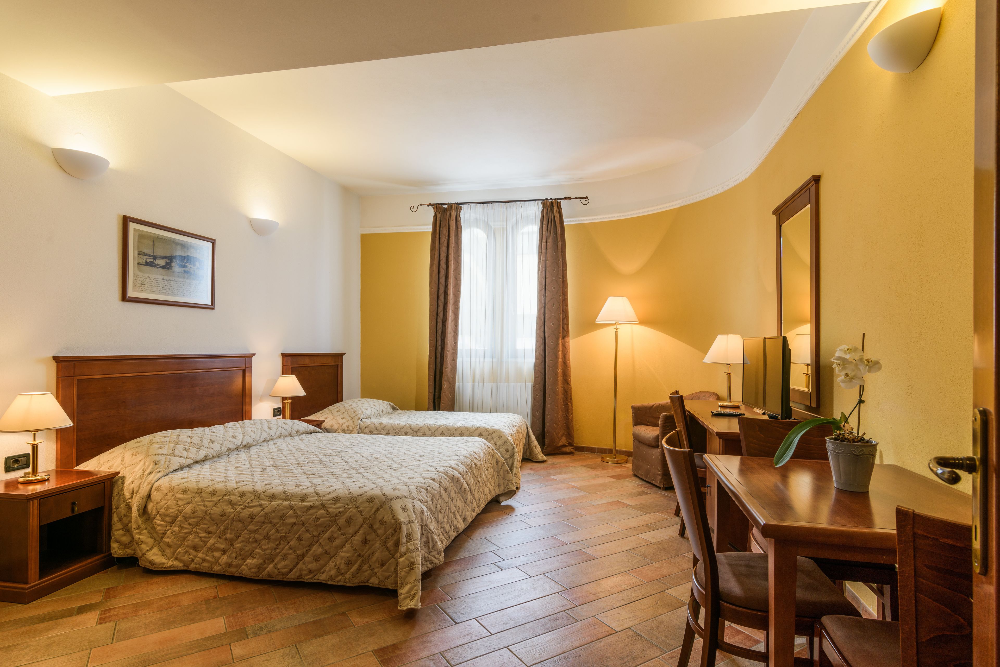 Ein Hotelzimmer mit zwei Betten, einem Schreibtisch, einem Stuhl, einer Lampe, einem Spiegel und einer Zimmerpflanze.