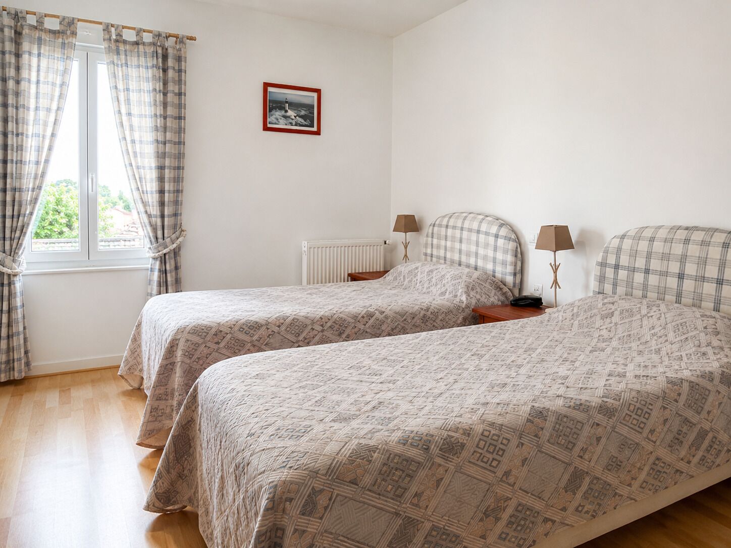 A bedroom with two beds, a window with curtains, a radiator, and a framed picture on the wall.