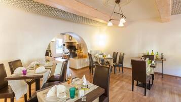 A dining area with tables set for guests, a kitchen in the background, and a wooden ceiling.