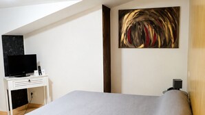 A bedroom with a bed, a desk with a television, a painting on the wall, and a lamp on a side table. - Auberge La Crémaillère (Villandraut)