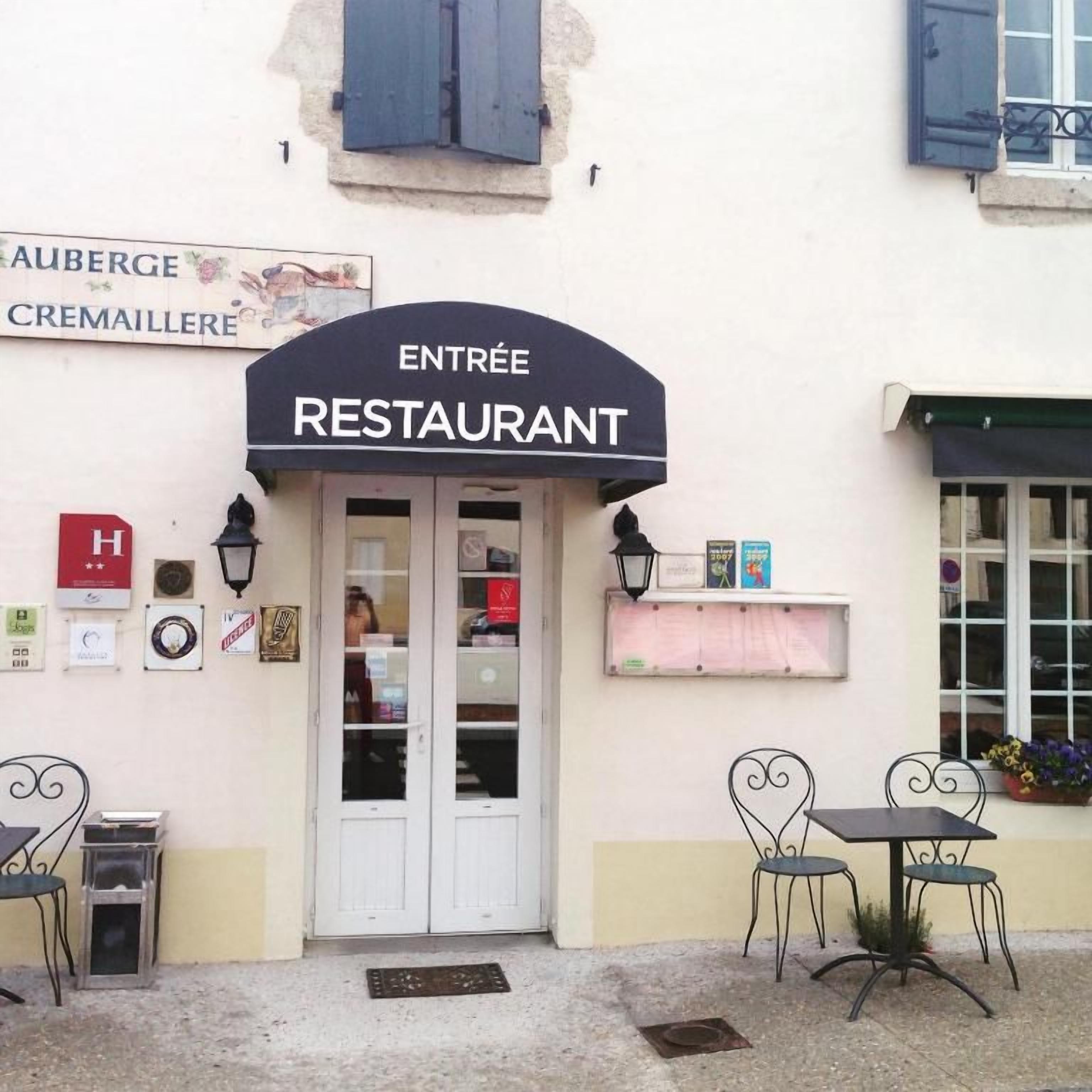 Entrada de un restaurante con un letrero, dos sillas, una mesa y un pequeño espacio para sentarse al aire libre.