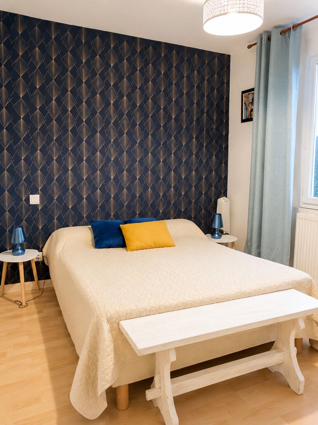 A bedroom with a bed, bedside tables, a bench, a patterned wall, and a window with curtains.