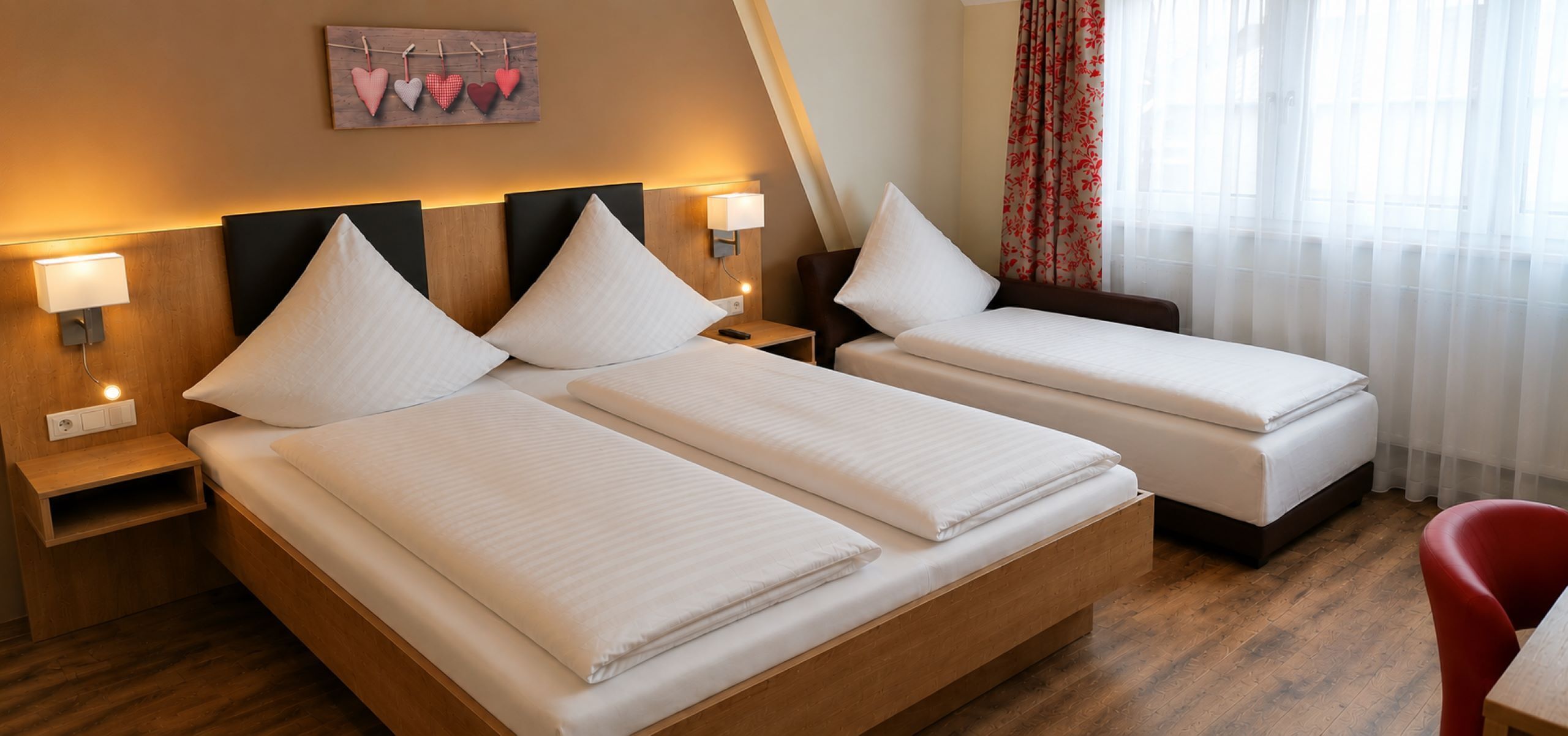 Habitación de hotel con dos camas, cabeceros de madera, lámparas de noche, mesitas de noche, una silla roja y una ventana con cortinas traslúcidas.