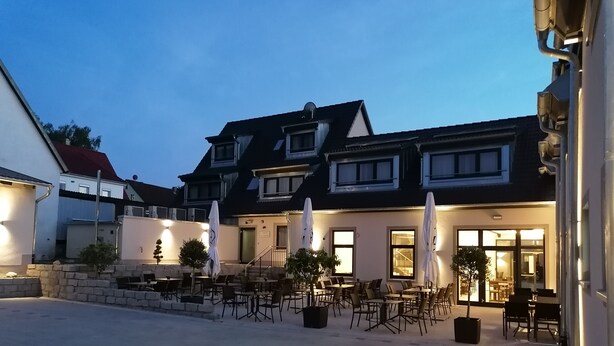 An outdoor seating area with tables and chairs, a stone wall, and a building with multiple windows and a staircase. - Landgasthof Niebler (Adelsdorf)