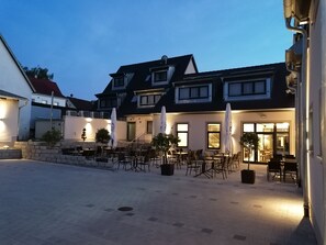 Ein Sitzbereich im Freien mit Tischen und Stühlen, eine Steinmauer und ein Gebäude mit mehreren Fenstern und einer Treppe.
