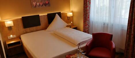 A hotel room with a bed, bedside tables, a red chair, a wall-mounted air conditioner, and a window with sheer curtains.