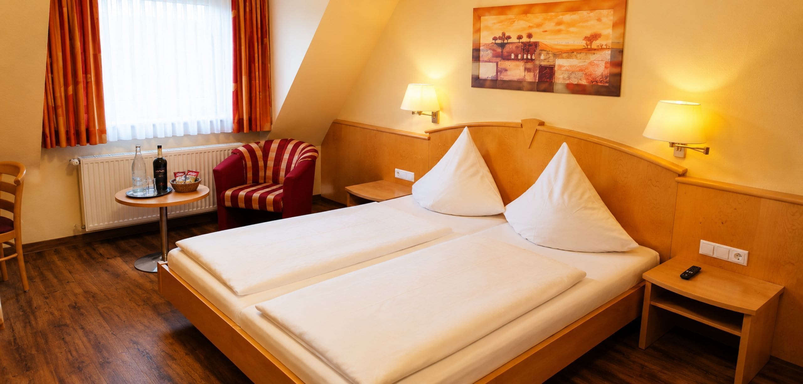 A hotel room with a bed, two bedside tables, a chair, a small table with a bottle and snacks, and a painting on the wall.