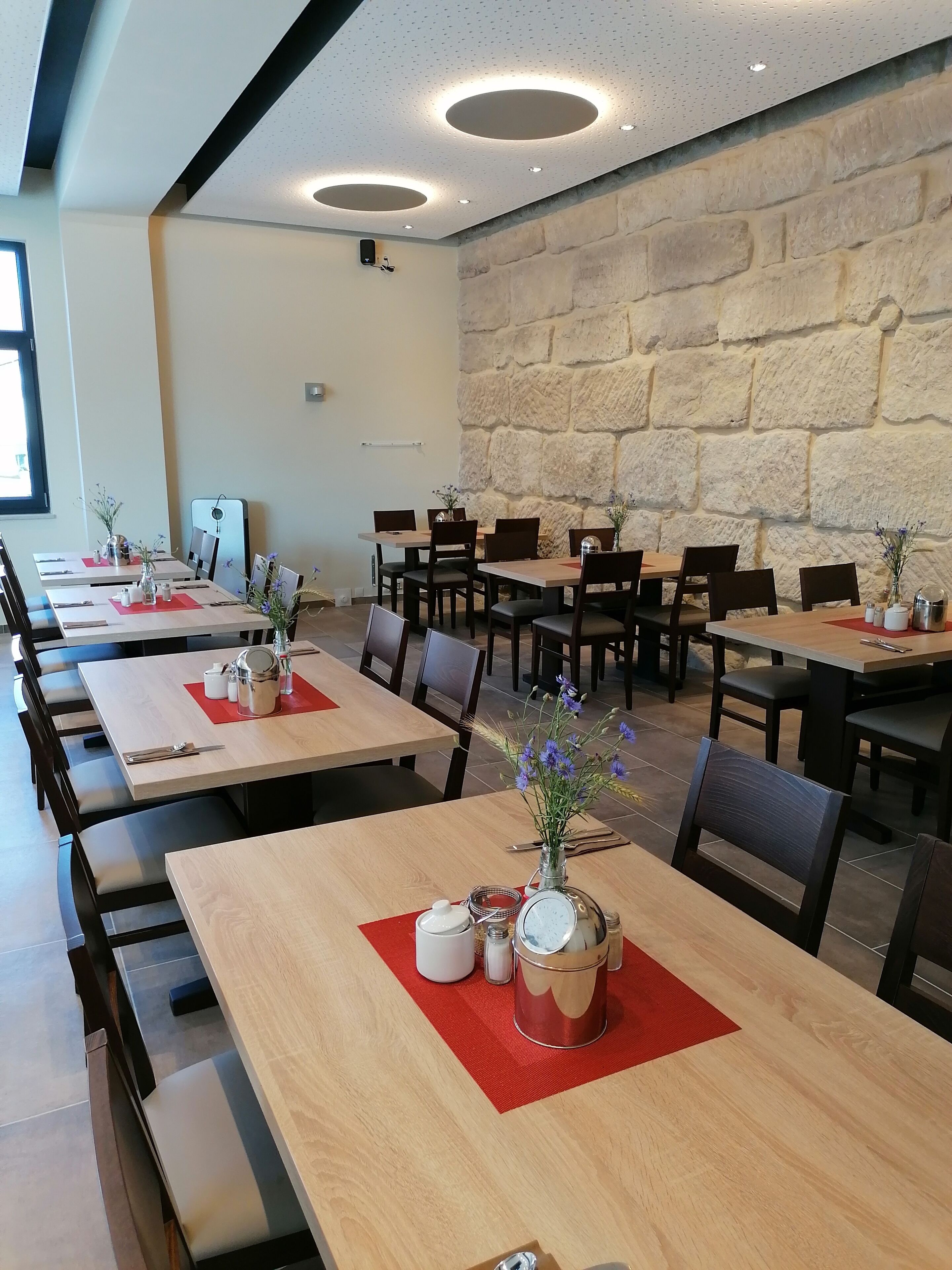 Un restaurante con varias mesas, cada una con manteles individuales rojos, teteras blancas y jarrones con flores. Las paredes presentan un diseño de piedra, y el techo tiene luces circulares.
