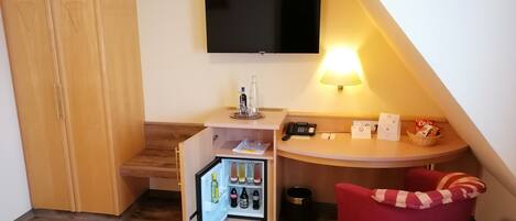 A compact room with a wooden wardrobe, a small kitchenette with a fridge, a TV mounted on the wall, a desk with a lamp, and a red chair.