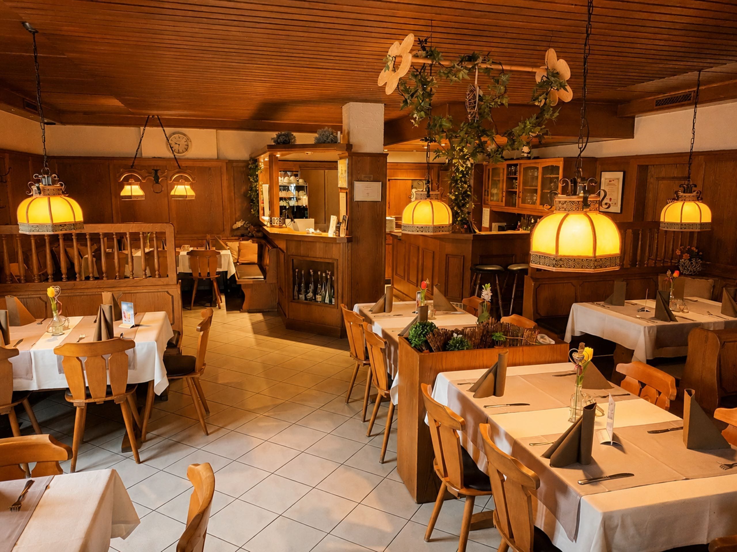 Un restaurante con interiores de madera, mesas dispuestas con manteles blancos y adornos florales colgantes.