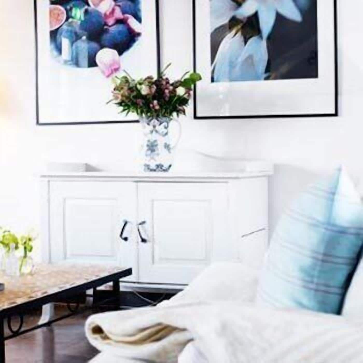 A room with a white cabinet, a table with a vase of flowers, and a blue and white striped pillow on a white sofa.