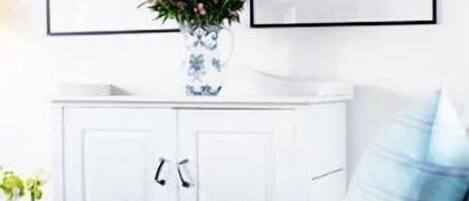 Una habitación con un armario blanco, una mesa con un jarrón de flores y un cojín azul y blanco a rayas sobre un sofá blanco.