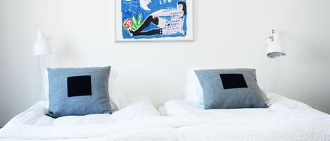 Una cama bien hecha con ropa blanca y dos almohadas, un cartel enmarcado en la pared y una lámpara blanca al lado de la cama.