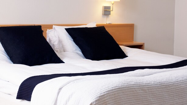 A neatly made bed with white and navy bedding, a wooden headboard, and a wall-mounted lamp.